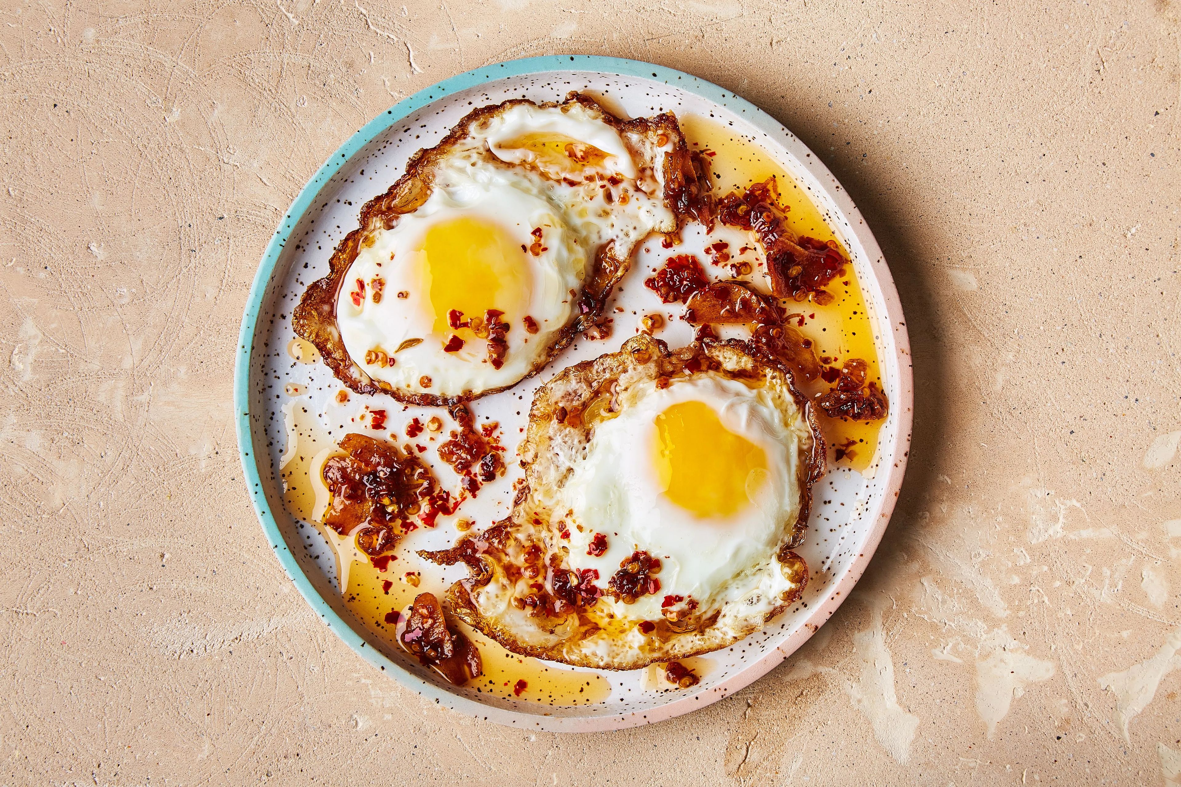 Two sunny-side-up eggs with crispy edges, drizzled with Silas & Co. Bushveld Heat chili oil and garlic crisps, served on a speckled plate against a textured beige background.